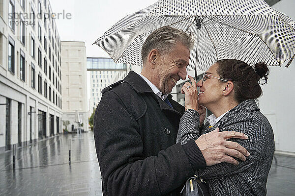 Verspielte Geschäftsfrau mit älterem Geschäftsmann unter Regenschirm in der Stadt während der Regenzeit stehend