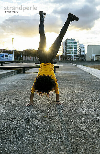 Junge Frau mit Afro-Haar macht Handstand auf der Straße gegen den Himmel bei Sonnenuntergang