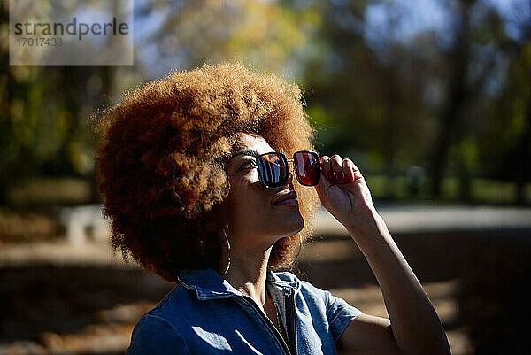 Schöne Frau schaut nach oben und hält eine Sonnenbrille in einem öffentlichen Park an einem sonnigen Tag