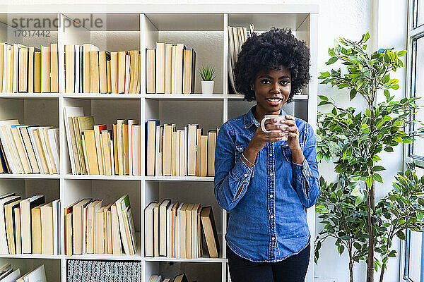 Lächelnde junge Frau trinkt Kaffee  während sie zu Hause am Bücherregal steht