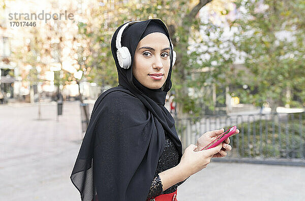 Porträt einer jungen schönen Frau mit Hijab und Kopfhörern  die ein Smartphone benutzt