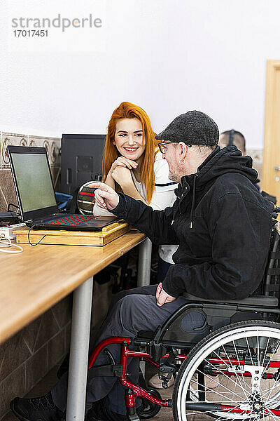 Lächelnde Betreuerin im Gespräch mit einem behinderten Mann am Laptop im Pflegeheim
