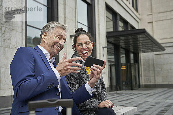 Fröhliche männliche und weibliche Unternehmer teilen sich ein Smartphone  während sie an einem Gebäude sitzen