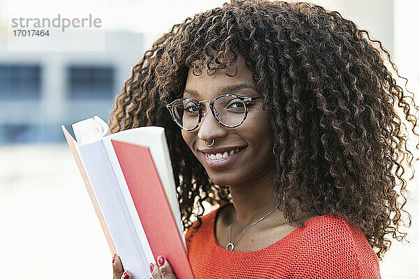 Lächelnde junge Frau mit lockigem Haar  die ein Buch hält