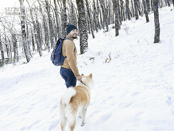Lächelnder junger Mann  der wegschaut  während er mit einem Akita-Hund im verschneiten Land steht