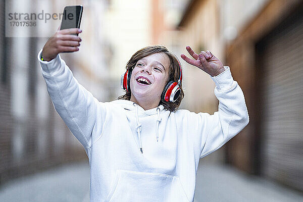 Lächelnder Junge mit Kopfhörern  der eine Friedensgeste zeigt  während er ein Selfie mit seinem Mobiltelefon im Freien macht