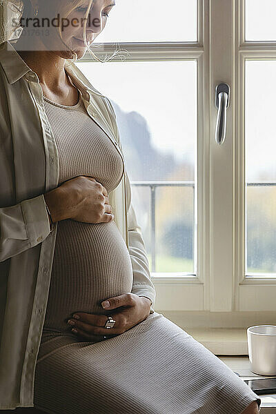 Schwangere Frau mit Händen auf dem Bauch sitzt am Fenster zu Hause