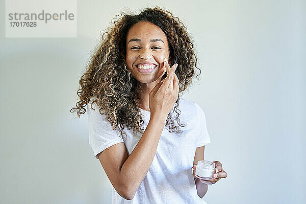 Fröhliche junge Frau  die Gesichtscreme vor einer weißen Wand aufträgt