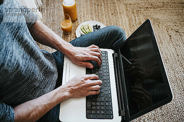 Mann benutzt Laptop  während er beim Frühstück zu Hause auf dem Boden sitzt