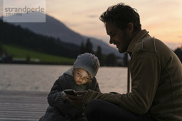 Vater und kleine Tochter spielen in der Abenddämmerung mit ihrem Smartphone auf einem Steg am Seeufer