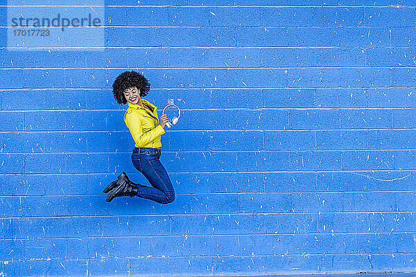 Fröhliche junge Frau hält Kopfhörer  während sie gegen eine blaue Wand springt