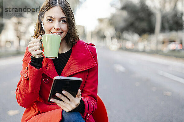 Modische junge Frau  die auf der Straße einen Kaffee trinkt und ein digitales Tablet hält