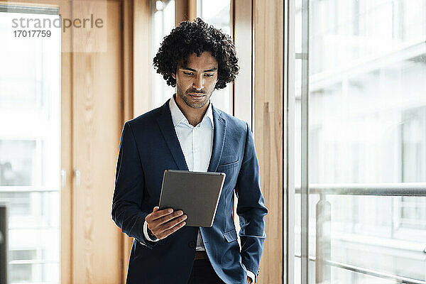 Selbstbewusster junger männlicher Unternehmer mit lockigem schwarzem Haar  der im Büro ein digitales Tablet benutzt