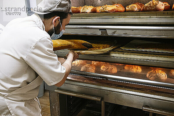Männlicher Bäcker mit Pizzaschieber bei der Kontrolle von Brot im Ofen in einer Bäckerei während COVID-19