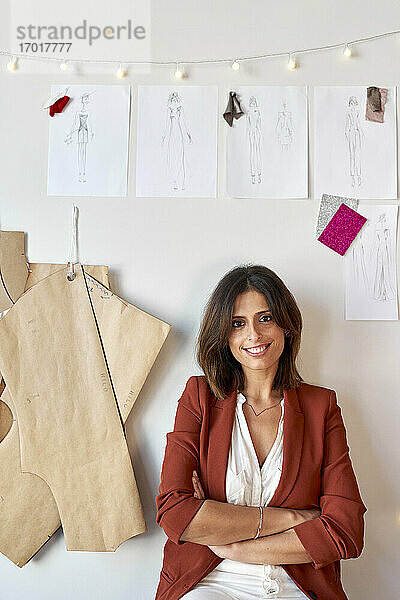 Selbstbewusst lächelnde Modedesignerin  die mit verschränkten Armen unter ihren Entwürfen und Mustern an einer weißen Wand im Atelier sitzt