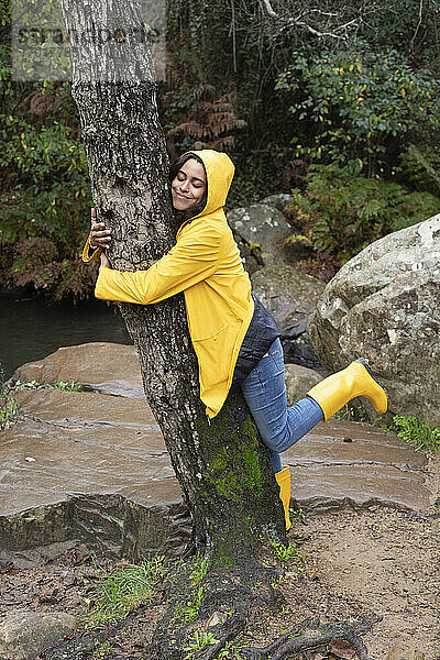 Glückliche Wanderin in gelbem Regenmantel umarmt einen Baum im Wald