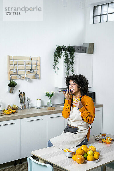 Lächelnde Frau mit Orangensaft  die mit ihrem Handy telefoniert  während sie zu Hause auf einem Tisch mit Obst sitzt