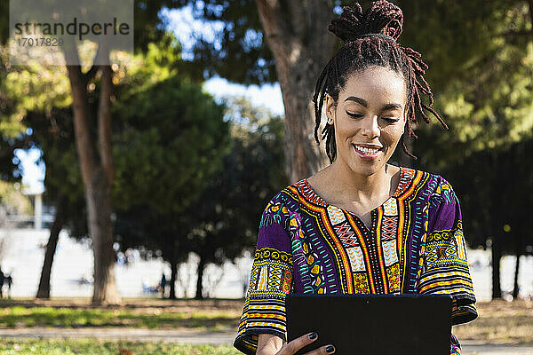 Schöne jamaikanische Frau mit Dreadlocks  die ein digitales Tablet im Park an einem sonnigen Tag benutzt