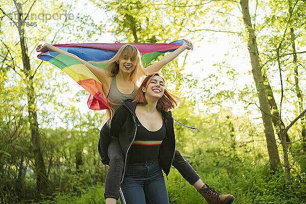 Glückliche Freundin  die eine Frau mit Regenbogenfahne huckepack nimmt  während sie im Wald steht