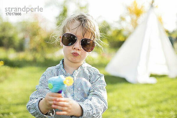 Mädchen mit Sonnenbrille  das mit einer Seifenblasenpistole spielt  während es im Garten steht