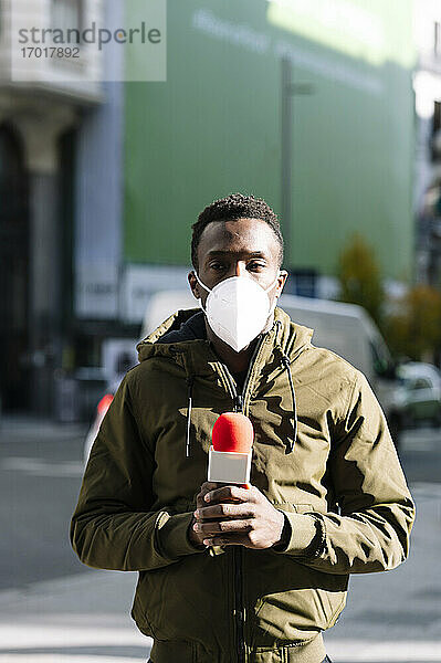 Männlicher Fernsehreporter mit Gesichtsmaske  der ein Mikrofon hält  während er auf einer Straße in der Stadt steht