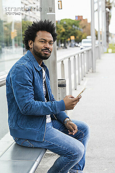 Selbstbewusster Mann mit Afro-Haar  der ein Mobiltelefon benutzt  während er auf einem Sitz an einer Bushaltestelle sitzt