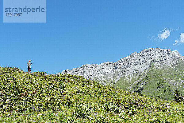 Mittlerer erwachsener Mann  der ein Mobiltelefon benutzt  während er auf einer Wiese am Col des Aravis steht  Haute-Savoie  gegen den Himmel  Frankreich