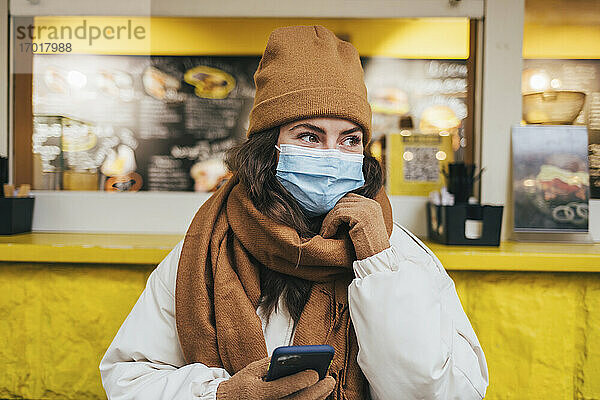 Junge Frau mit Hand am Kinn träumt  während sie ihr Smartphone in einem Café während der COVID-19 hält