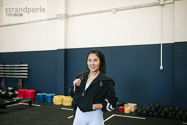 Lächelnde Sportlerin mit Händen in den Taschen  die einen Rucksack im Fitnessstudio trägt