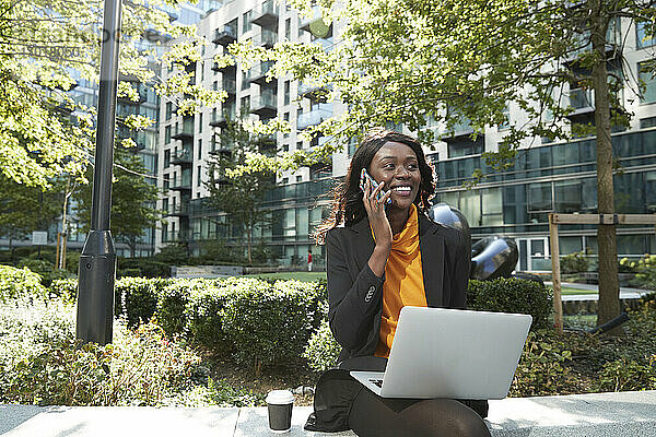 Lächelnde Geschäftsfrau mit Laptop im Gespräch am Telefon im Büropark