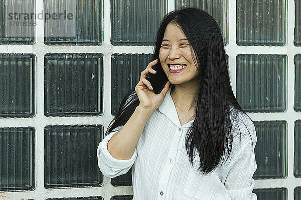 Fröhliche Frau  die mit einem Mobiltelefon spricht  während sie an einer Glaswand steht