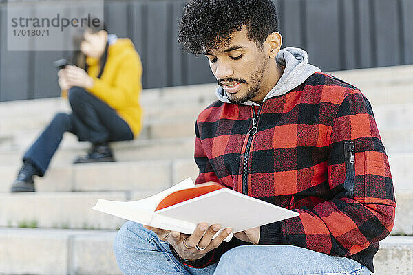 Junger Mann mit Kapuzenshirt liest ein Buch  während er auf einer Treppe neben einem Skateboard sitzt
