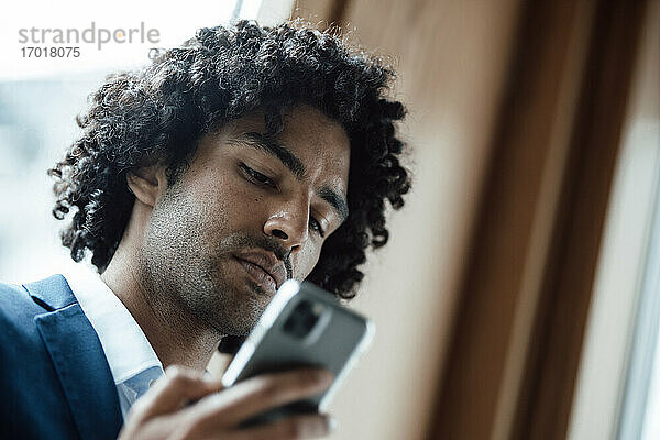 Junger männlicher Unternehmer mit lockigem schwarzem Haar  der im Büro ein Smartphone benutzt