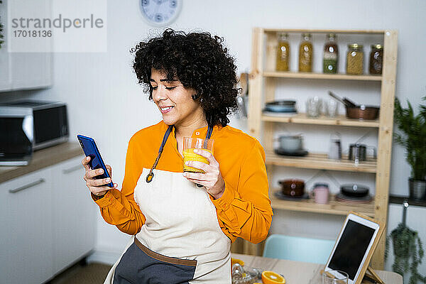 Junge Frau mit Orangensaft und Mobiltelefon  während sie zu Hause auf dem Tisch sitzt