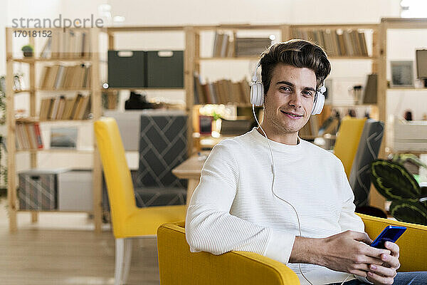 Lächelnder junger Mann hört Musik über Kopfhörer und hält ein Handy in seinem neuen Zuhause