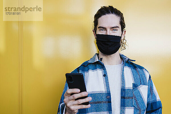 Junger Mann mit Schutzmaske und Mobiltelefon  der gegen eine gelbe Wand starrt