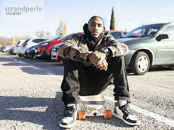 Junger Mann mit cooler Einstellung sitzt auf dem Skateboard auf einem Parkplatz an einem sonnigen Tag