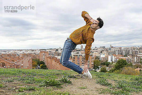 Flexibler junger Mann  der die Augen abschirmt  während er auf einem Hügel in der Stadt gegen den bewölkten Himmel akrobatische Übungen macht