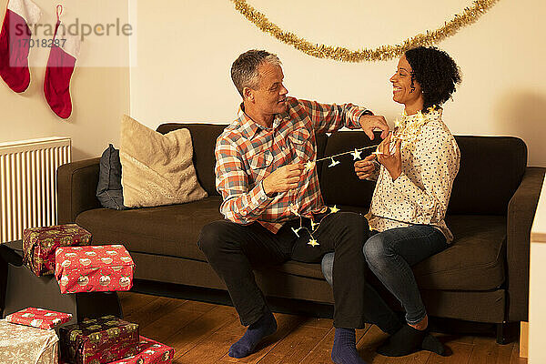 Fröhliches Paar spielt mit beleuchteten Weihnachtslichtern  während es zu Hause auf dem Sofa sitzt