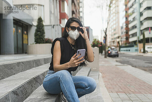 Frau mit Schutzmaske  die auf einer Treppe in der Stadt sitzt und ihr Smartphone benutzt