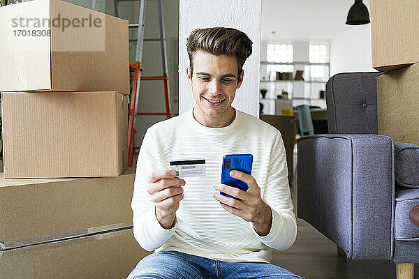 Lächelnder junger Mann  der beim Einzug in ein neues Haus mit dem Mobiltelefon und der Kreditkarte bezahlt