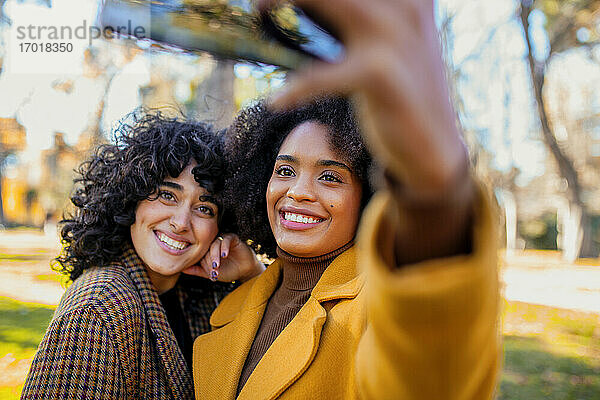 Lächelnde Frau  die ein Selfie mit einem Freund durch ein Smartphone macht  während sie im Park steht