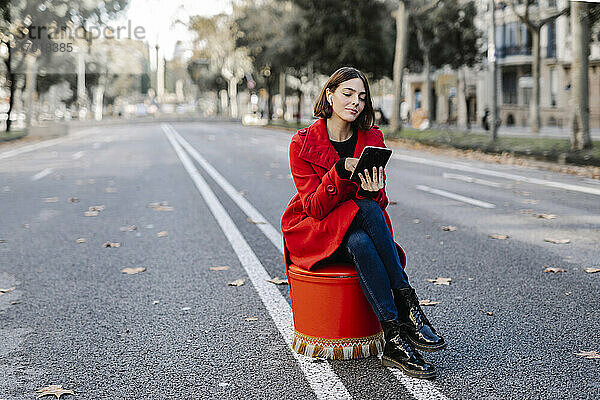 Junge Frau in roter Jacke hört Musik  während sie ein digitales Tablet benutzt  das auf einem Sitz auf der Straße steht