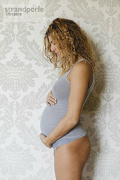 Schwangere Frau  die sich zu Hause an der Wand stehend um ihren Unterleib kümmert