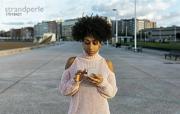 Junge Frau mit Afro-Haar  die ein Mobiltelefon benutzt  während sie auf der Straße steht