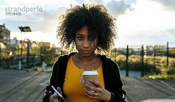 Junge Frau mit Afro-Haar hält Kaffee und Smartphone gegen den Himmel