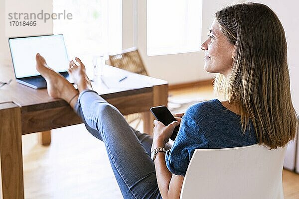 Lächelnde brünette Geschäftsfrau  die wegschaut  während sie mit Smartphone und hochgelegten Füßen am Tisch im Heimbüro sitzt