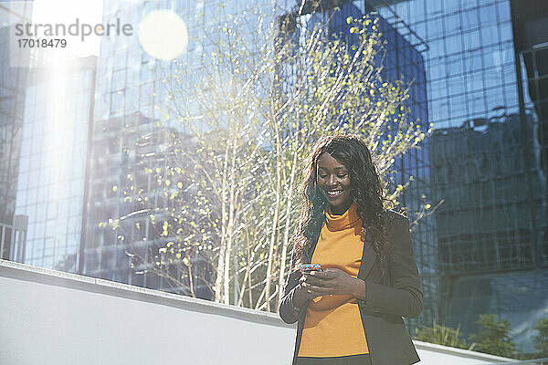 Glückliche Geschäftsfrau  die ihr Smartphone an einem sonnigen Tag vor einem Bürogebäude benutzt