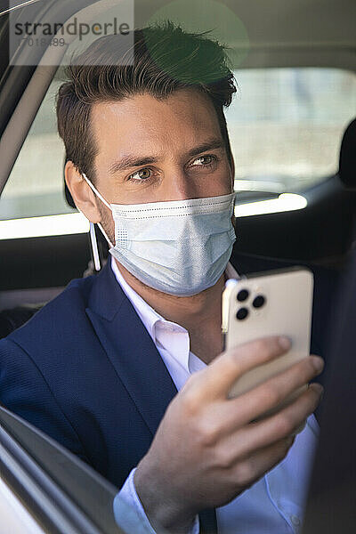 Nahaufnahme eines Geschäftsmannes mit Maske  der im Taxi ein Smartphone benutzt  gesehen durch ein Fenster