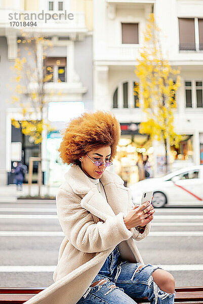 Frau mit afroblondem Haar benutzt ihr Smartphone  während sie auf einer Bank in der Stadt sitzt